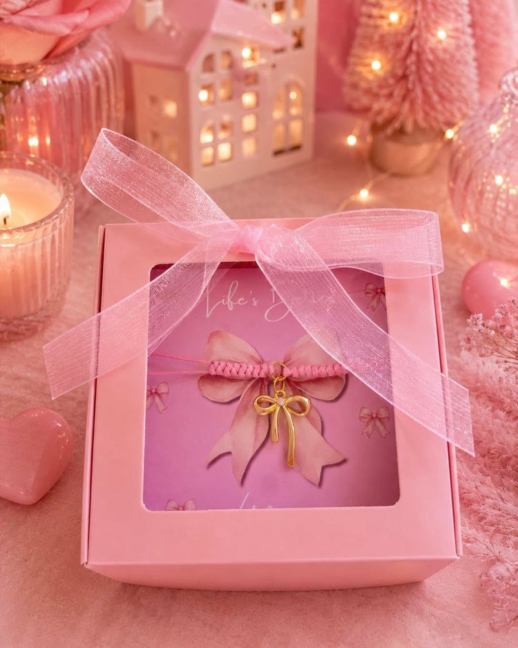 A wooden round decoration with a pink bow illustration and a sheer pink ribbon bow, styled on a pink fabric background with flowers, a candle, and a small heart-shaped object.