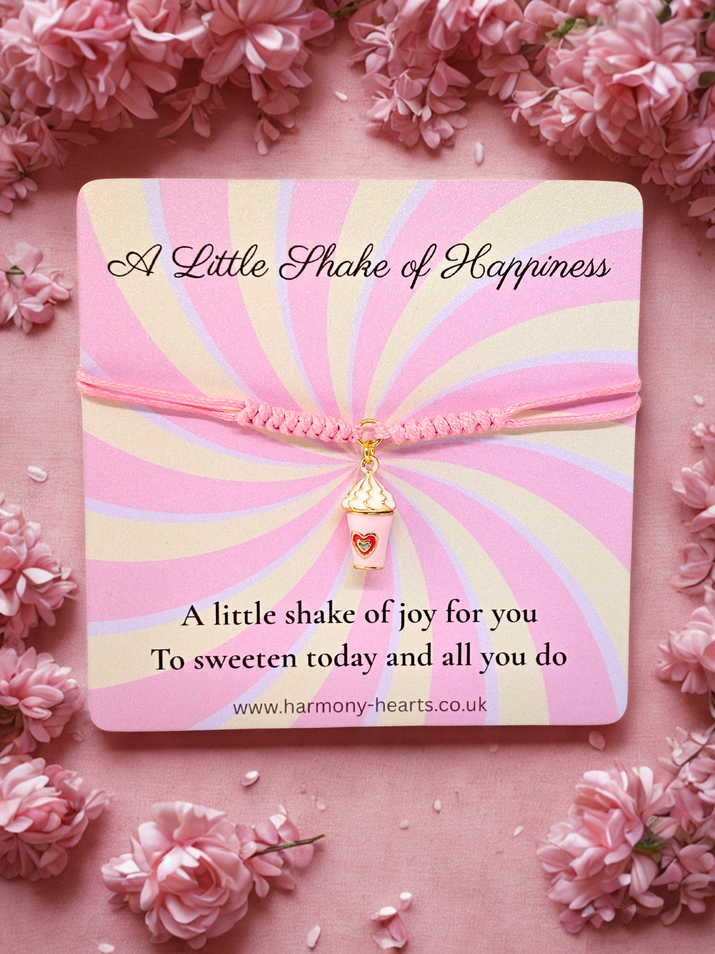 Pink and gold striped milkshake charm bracelet on 'A Little Shake of Happiness' branded card, surrounded by pink flowers.