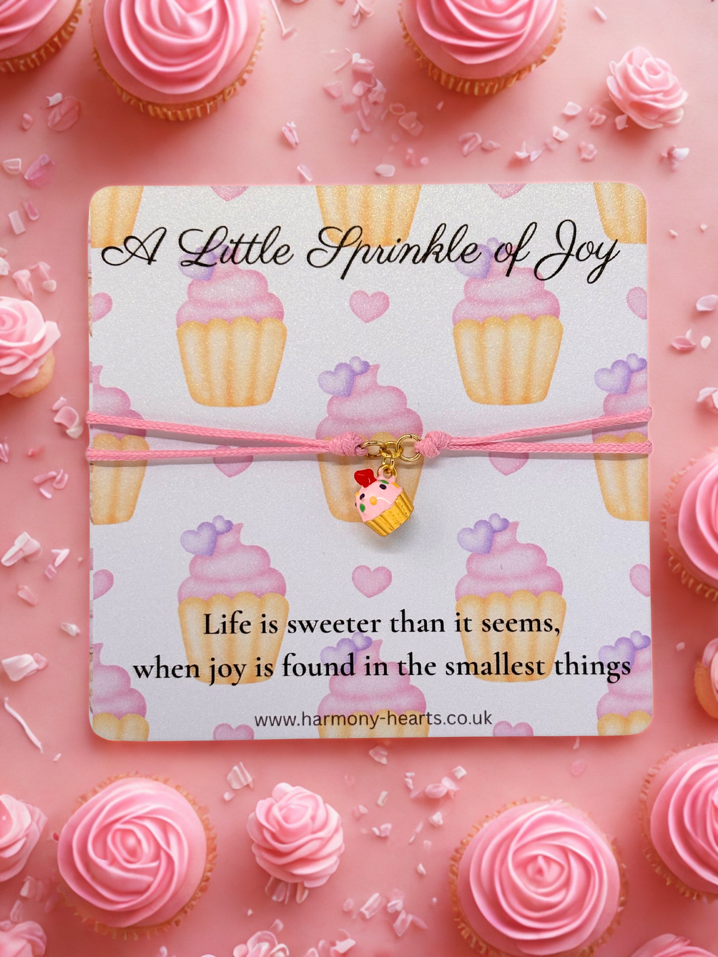 Pink bracelet with cupcake charm on a card with 'A Little Sprinkle of Joy' message, surrounded by pink cupcakes and sprinkles.
