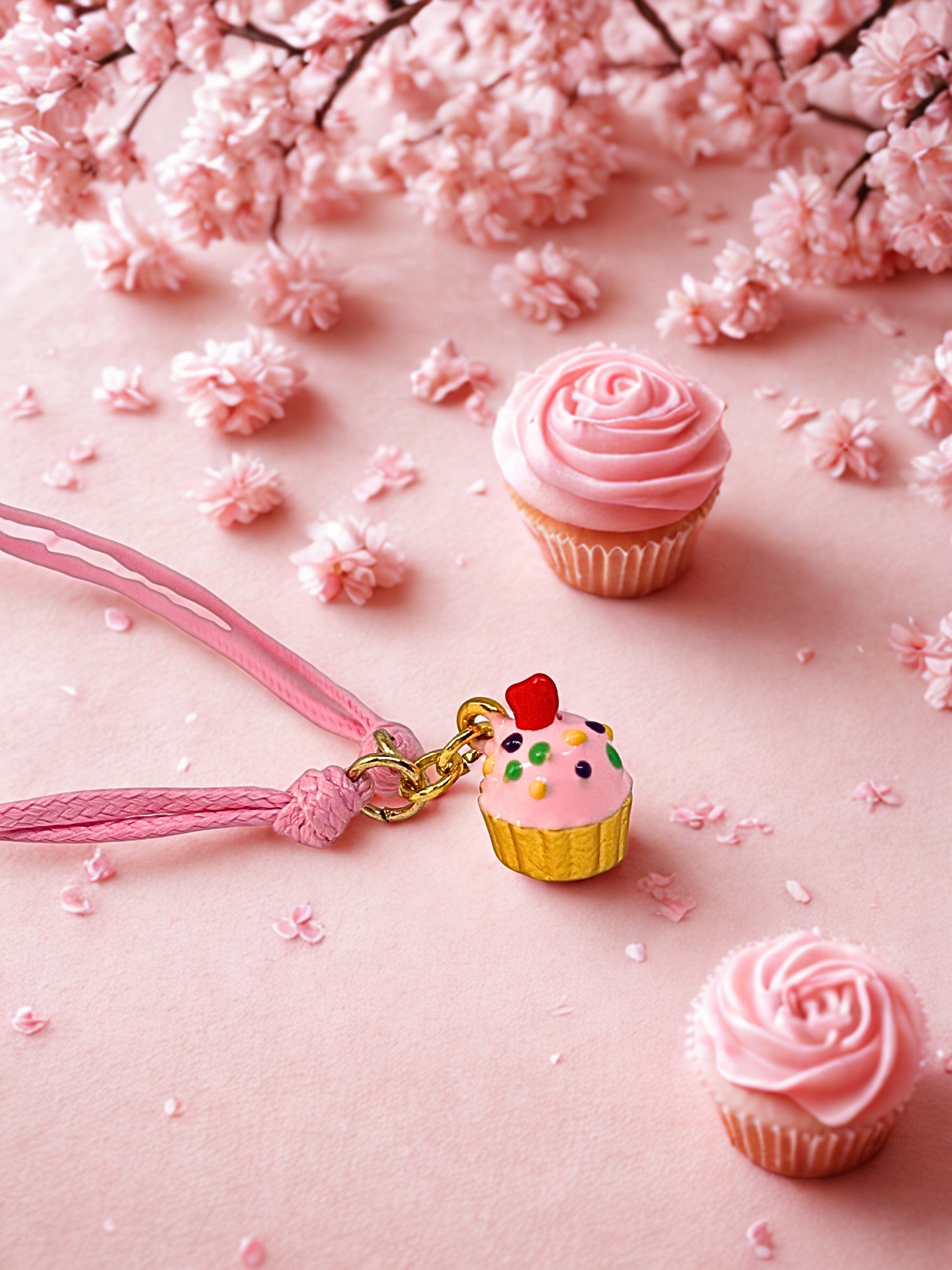 Cupcake-themed charm bracelet and small cupcakes on a pink background with cherry blossoms.