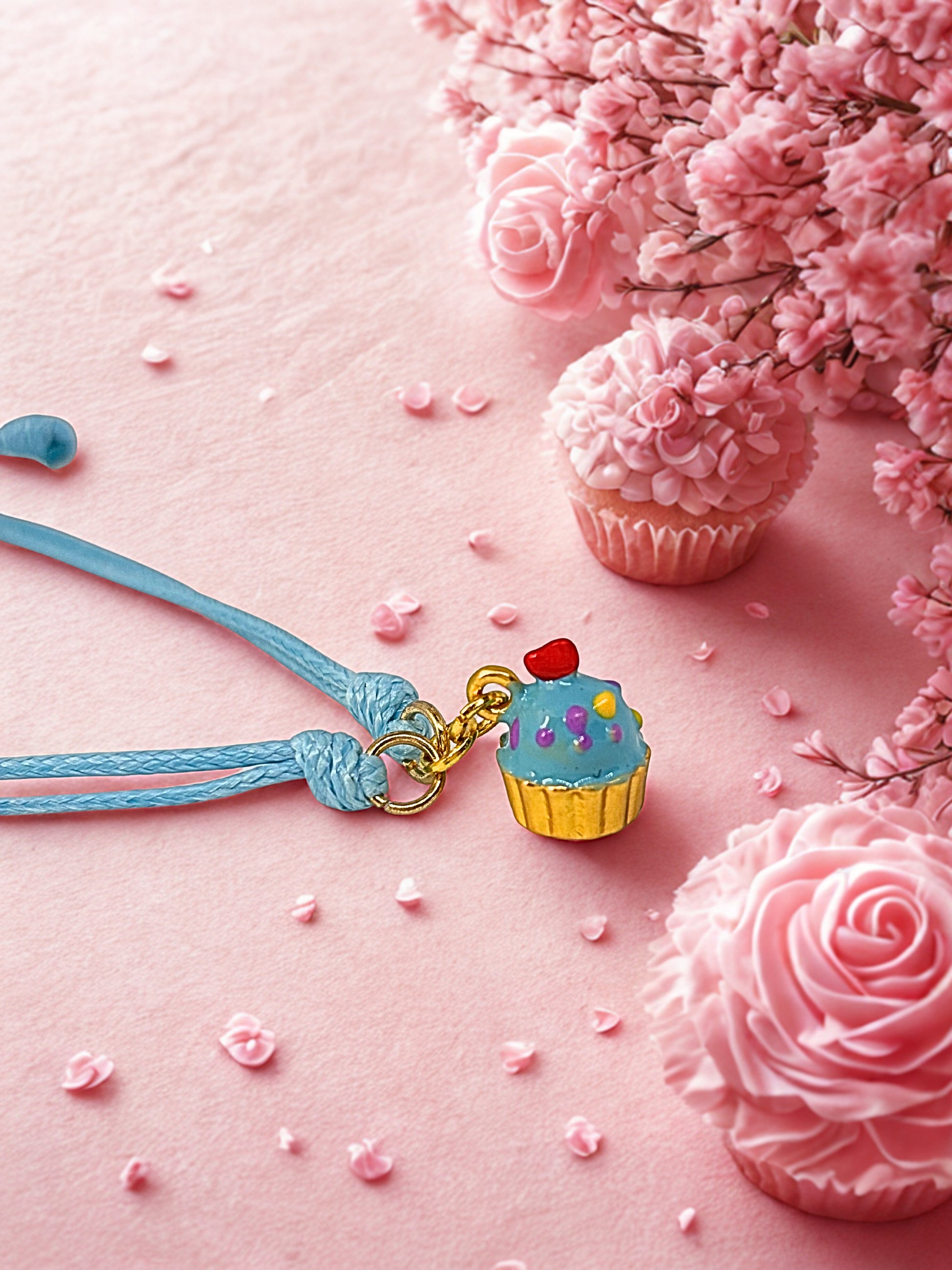 Cupcake-shaped charm bracelet on a blue cord with pink flowers and cupcakes on a pink background