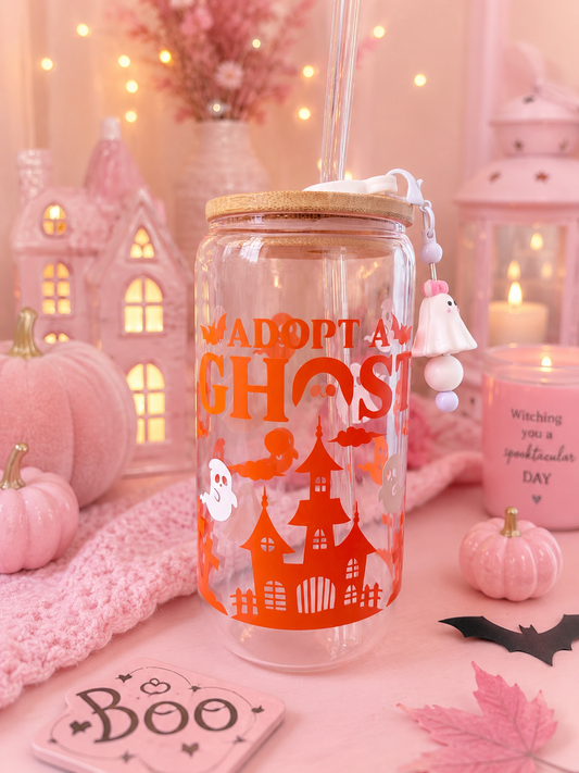 A clear glass tumbler with a bamboo lid and straw, decorated with an orange Halloween graphic that reads 'ADOPT A GHOST' with a haunted house and ghost illustration. The tumbler is styled in a cozy, pink-toned autumn setting with miniature pumpkins, a knitted scarf, a candle, and a 'Boo' coaster.