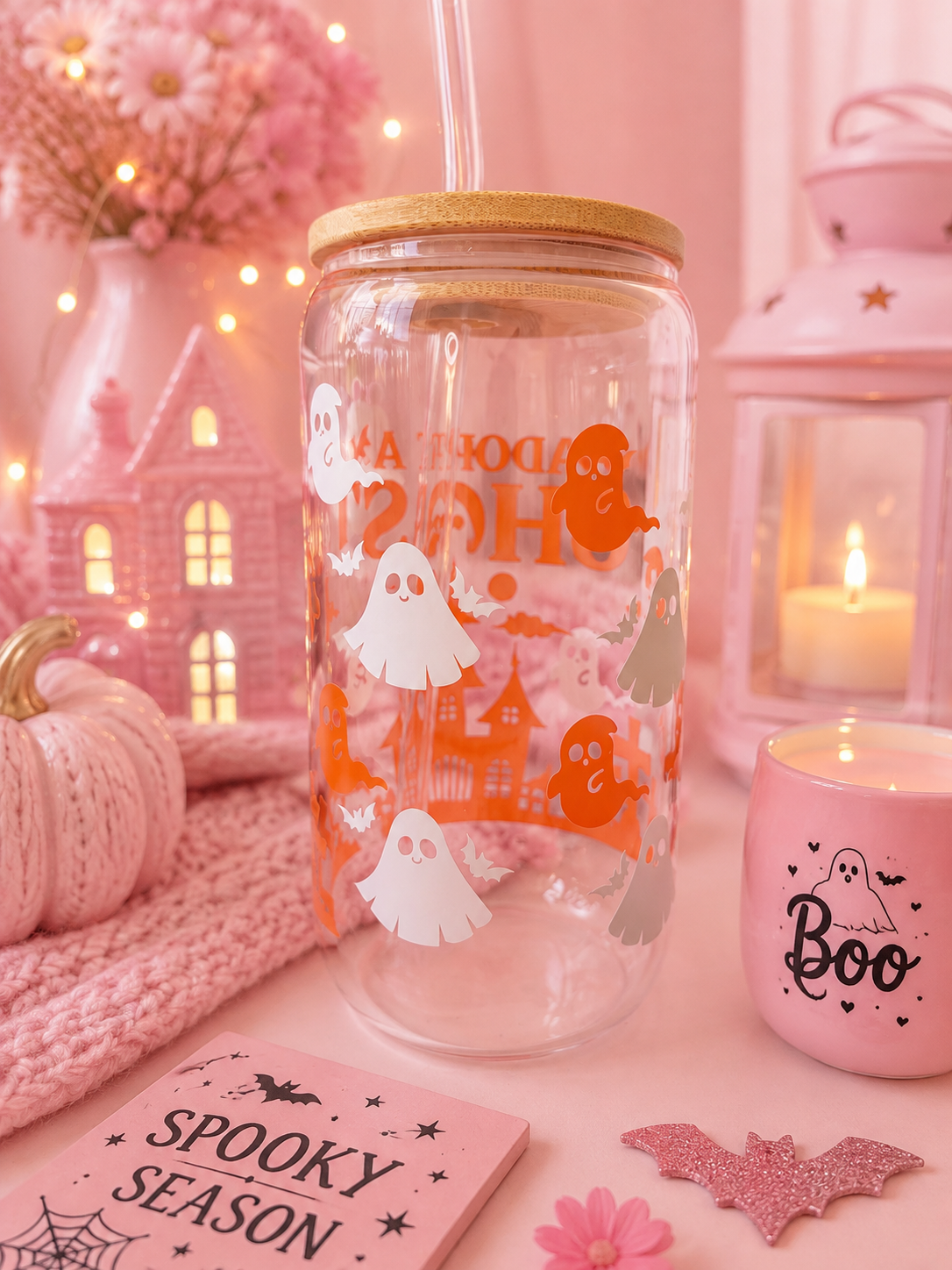 A clear glass mason jar tumbler with a bamboo lid and clear straw, decorated with orange and white Halloween graphics including ghosts, a haunted house. The tumbler is styled in a cozy pink autumn setting with a pink knit blanket, a pink candle, a small pumpkin, and a 'Spooky Season' card.