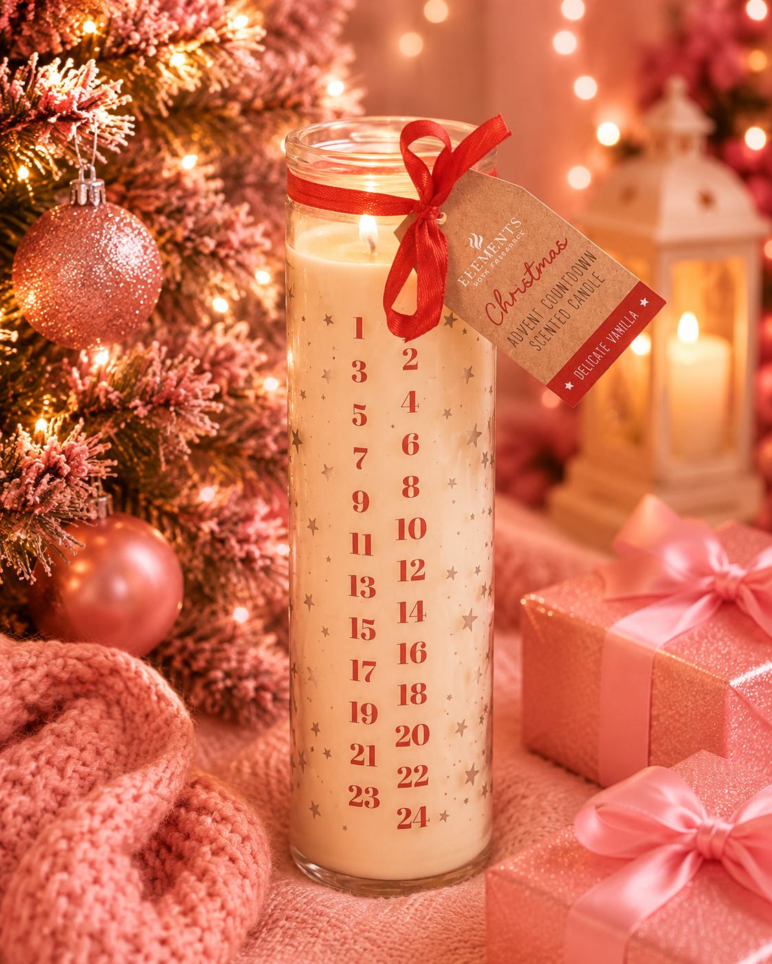 A tall glass jar candle with a red ribbon and hanging tag, decorated with a red numbered advent calendar print and small gold stars. The candle is styled on a table with pink wrapped gifts, a pink knit blanket, and a flocked Christmas tree with warm string lights in the background.