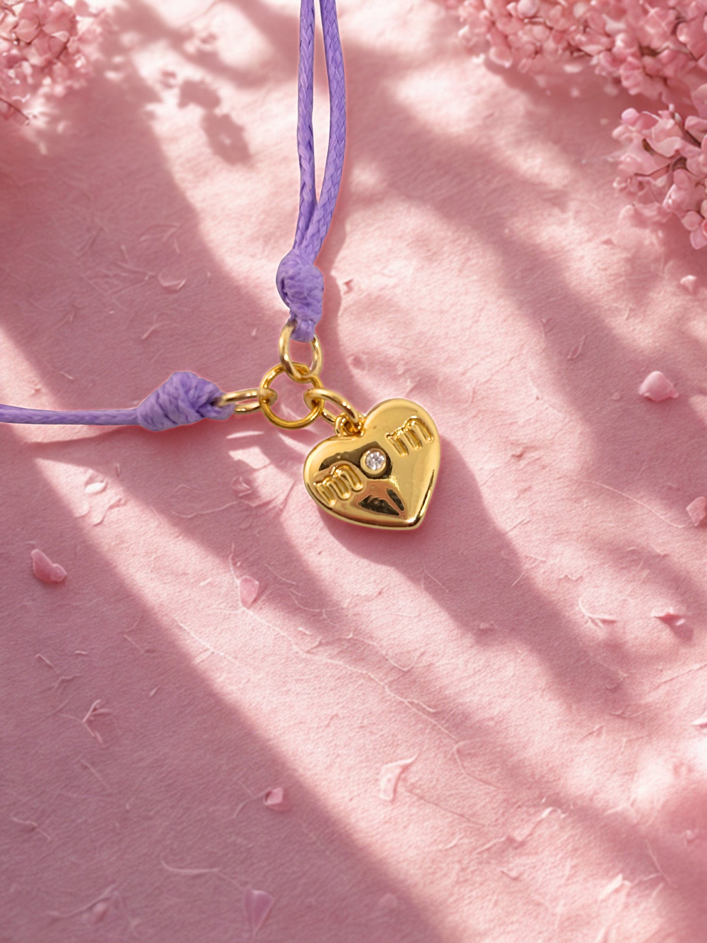 Gold heart-shaped charm bracelet on a purple cord against a pink background with cherry blossoms.