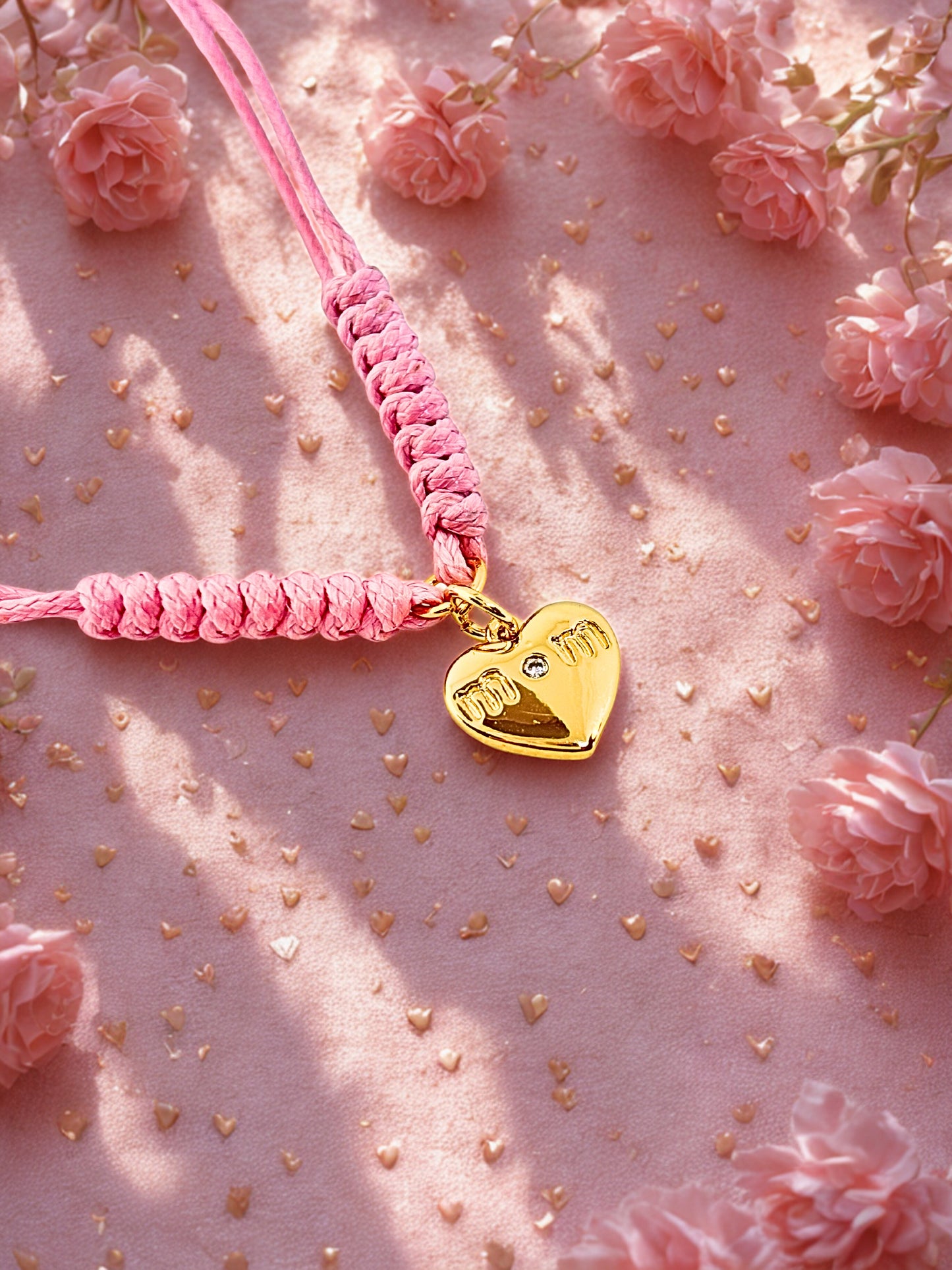 Pink braided cord bracelet with a gold heart-shaped charm on a pink background with flowers