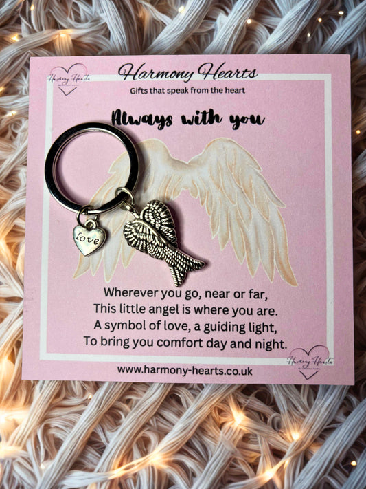 Keychain with angel wings and heart charm on a pink card with text, placed on a textured surface with string lights.