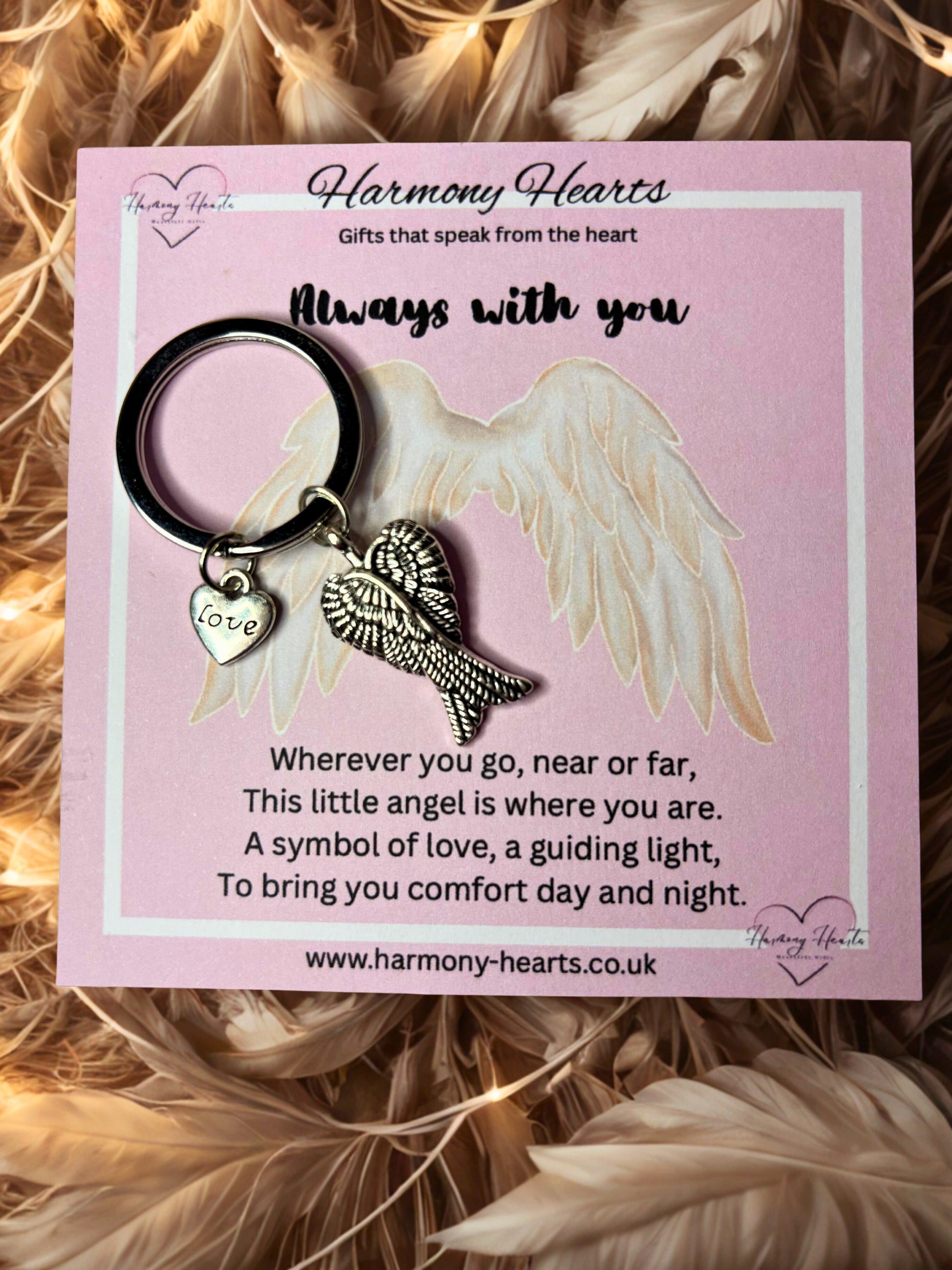Keychain with angel wings and heart on a pink card with text, surrounded by feathers.