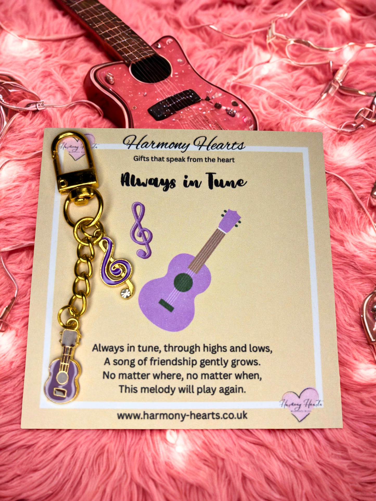 Purple ukulele-shaped keychain with gold chain on a 'Harmony Hearts' card against a pink textured background.
