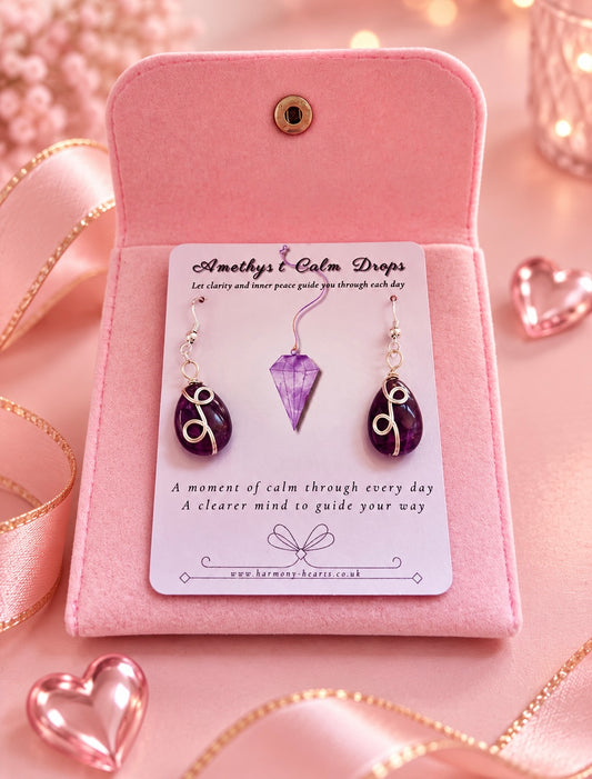 A pair of amethyst dangling earrings displayed on a pink presentation card inside a pink velvet case. The earrings have purple oval stones with white swirl details and silver hooks.