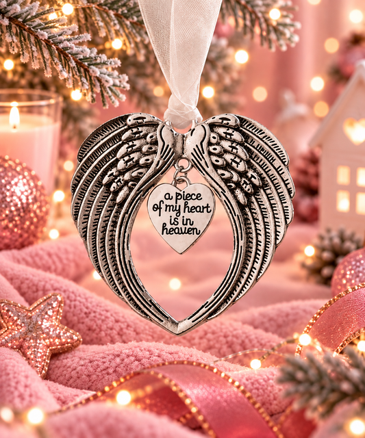 An ornate heart-shaped angel wing hanging ornament with a small heart charm that reads 'a piece of my heart is in heaven,' shown hanging in a cozy holiday setting with pink fabric, lights, and festive decorations.