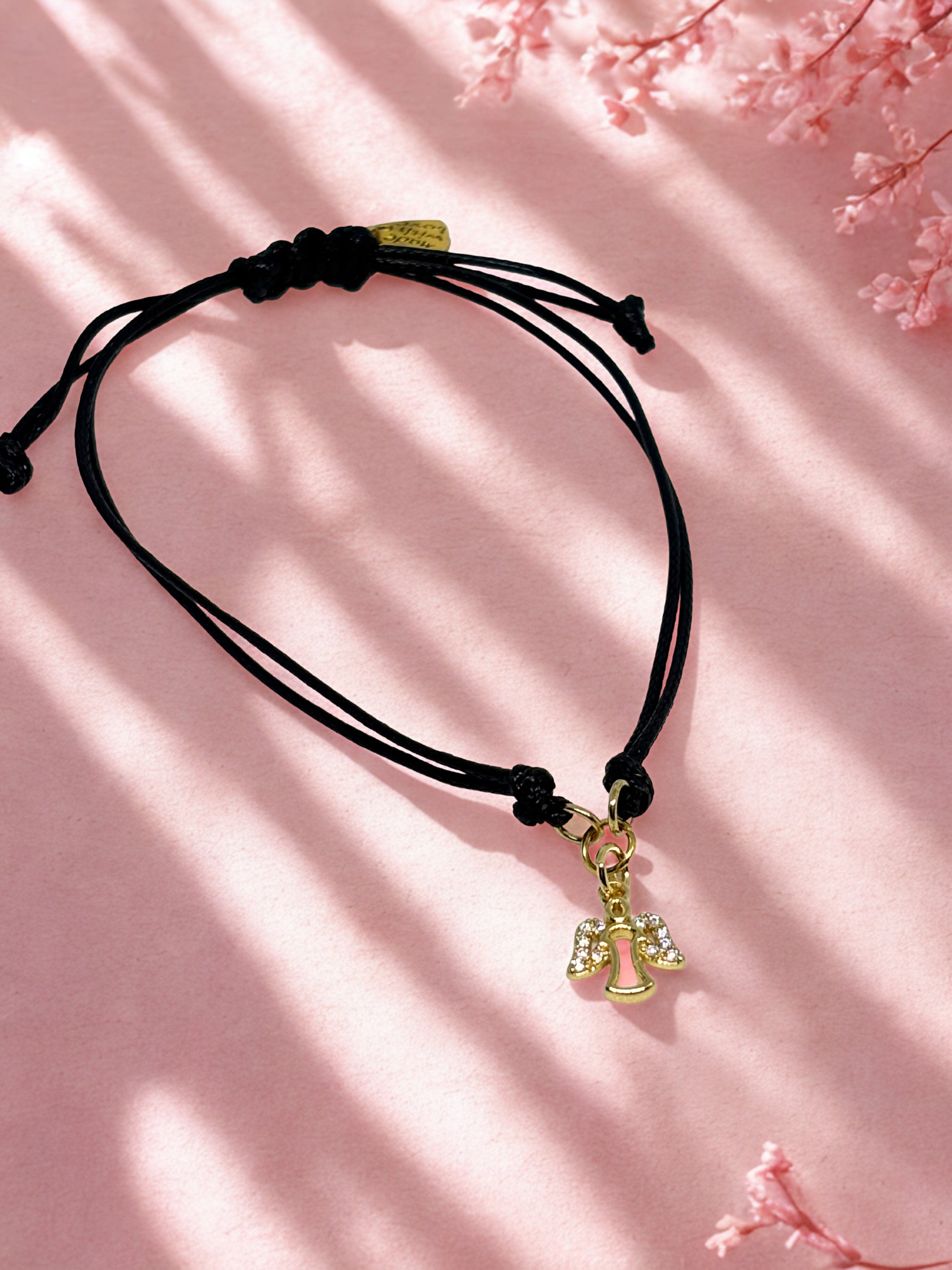 Black cord bracelet with gold angel charm on a pink background