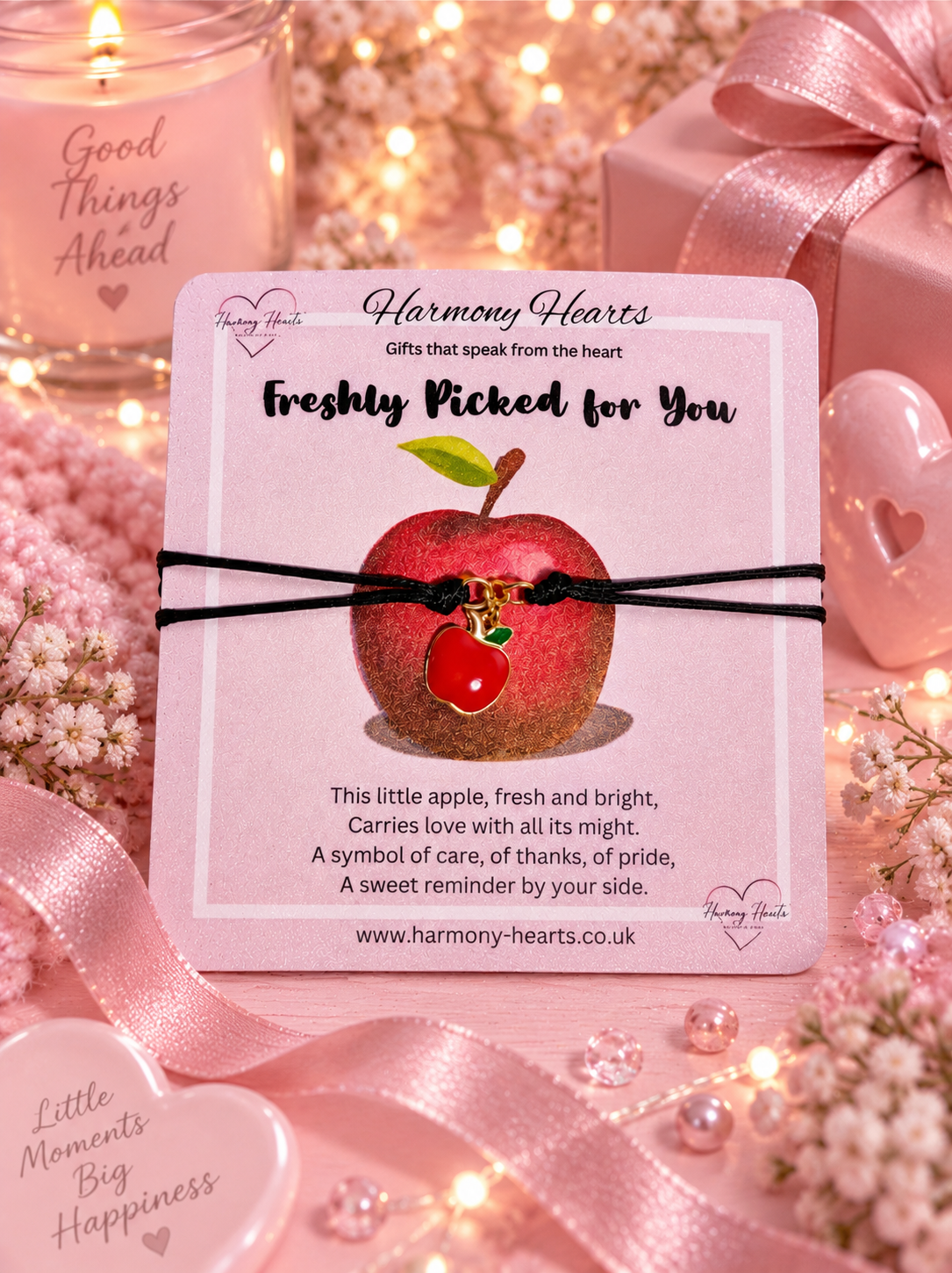 A Harmony Hearts card featuring a small apple charm on a black cord bracelet, surrounded by pink ribbons, hearts, and fairy lights.