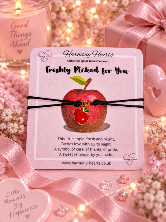 A Harmony Hearts card featuring a small apple charm on a black cord bracelet, surrounded by pink ribbons, hearts, and fairy lights.