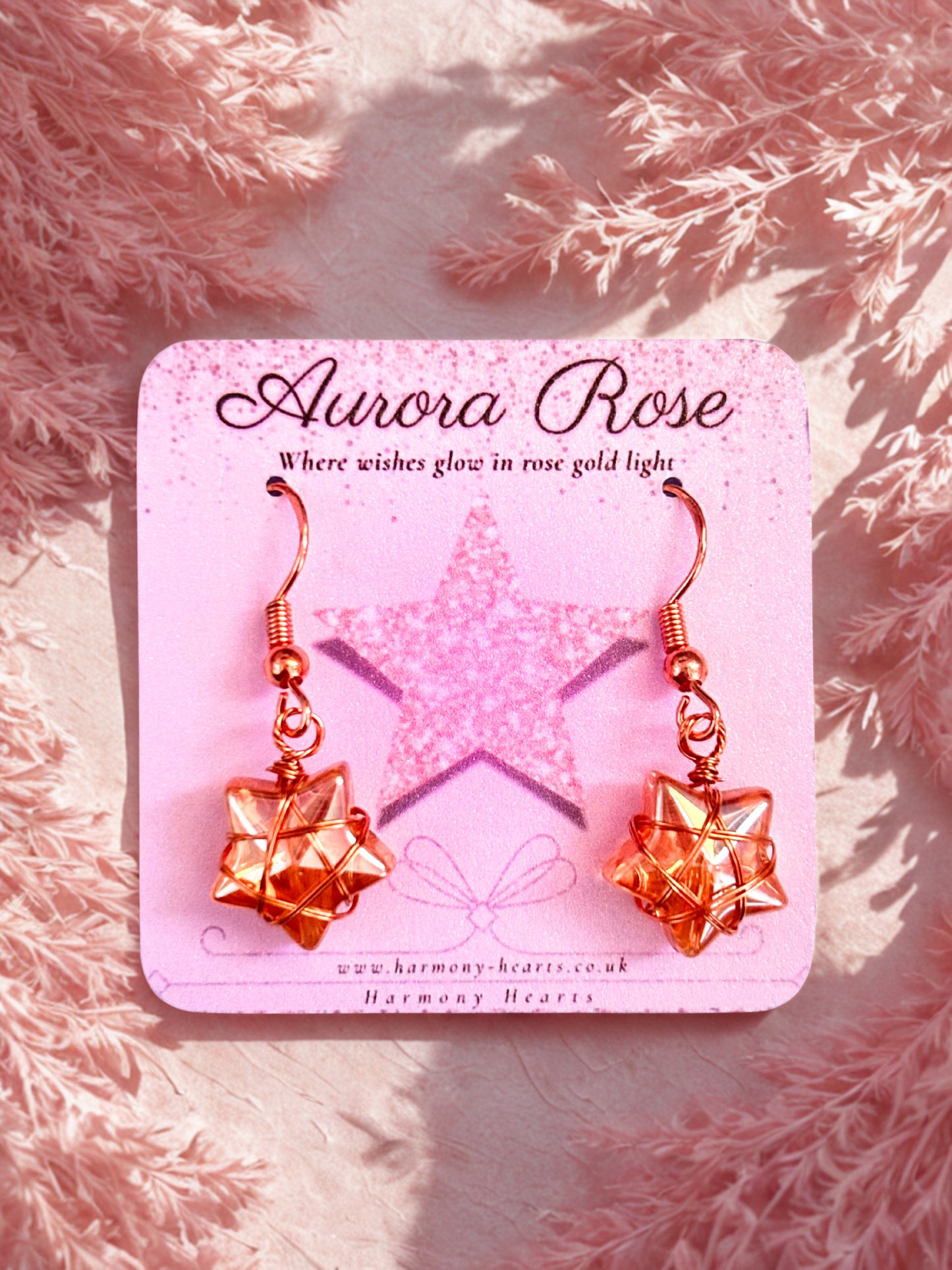Rose gold earrings on a pink 'Aurora Rose' branded card against a pink textured background.