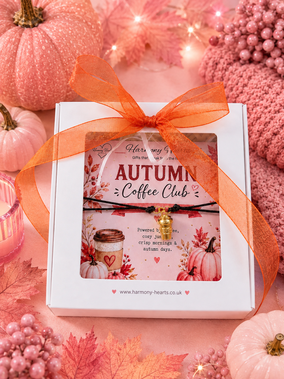 A white gift box tied with a sheer orange ribbon, featuring a window display of a 'Autumn Coffee Club' cord bracelet with a gold coffee cup charm. The box is styled on a pink background with autumn-themed props including pumpkins, leaves, and a pink knit hat.