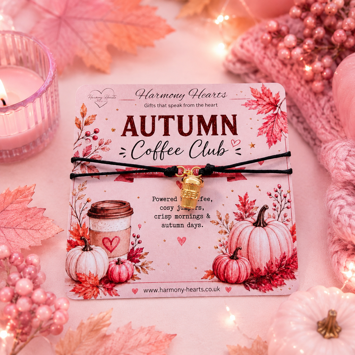 A black cord bracelet with a small gold coffee cup charm rests on a decorative printed card reading 'Autumn Coffee Club' from Harmony Hearts. The card is styled with autumn leaves, pumpkins, a candle, and pink knit textures in a soft, warm setting.
