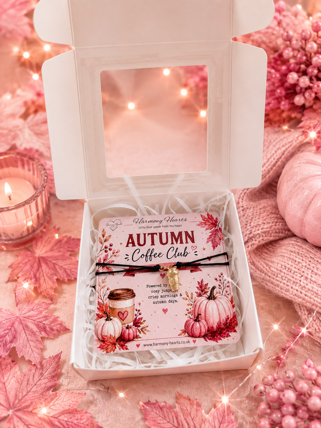An open white gift box filled with white paper shred holds a small printed card reading 'Autumn Coffee Club' with autumn illustrations. Presented with a black cord bracelet with gold coffee cup charm. The box is styled in a cozy pink setting with a candle, pink leaves, string lights, and a knitted pink scarf. 