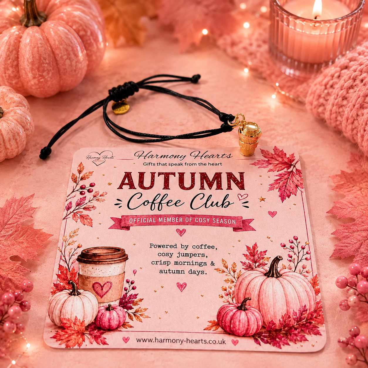 A square printed card for the Harmony Hearts Autumn Coffee Club is styled on a pink fabric surface with autumn leaves, small pumpkins, a candle, and a black cord bracelet with gold coffee cup charm.