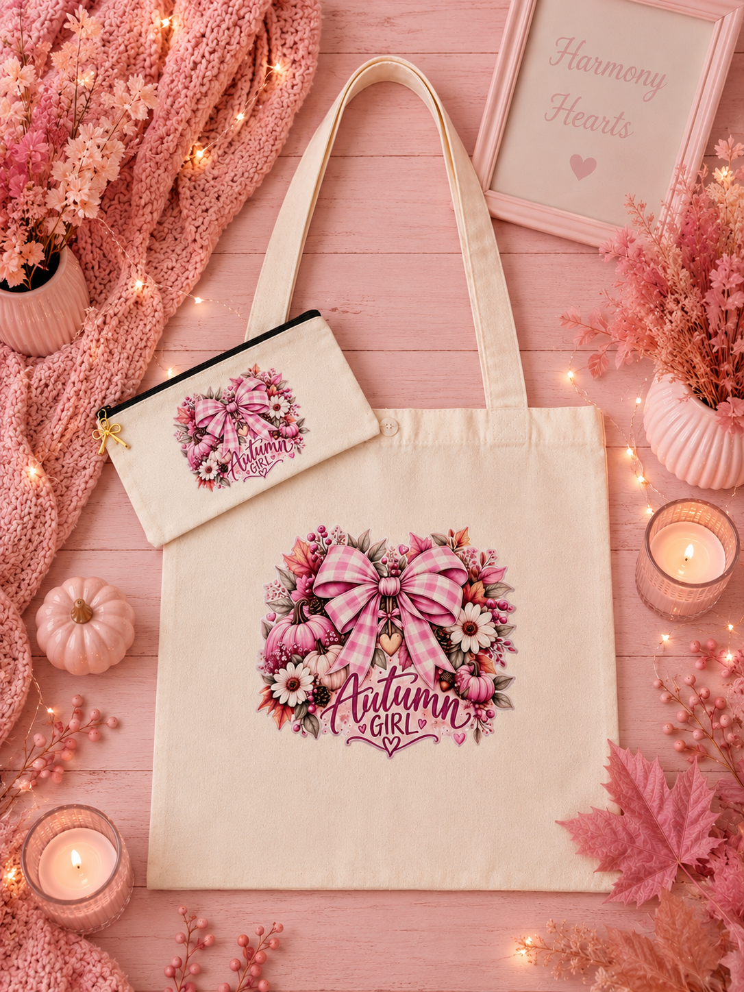 A cream canvas tote bag and matching zip pouch with a pink floral 'Autumn Girl' graphic, styled on a pink wood background with candles, dried flowers, a knit blanket, and fall leaves.