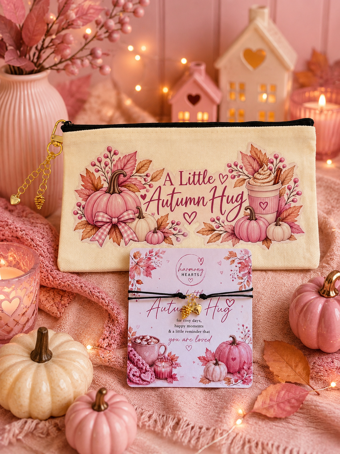 A cream fabric zip pouch printed with pink autumn pumpkins, leaves, and the phrase 'A Little Autumn Hug' is displayed with a matching greeting card with black cord bracelet attached. The scene is styled with pink knit blankets, small decorative pumpkins, string lights, and a cozy house-shaped candle holder in the background.