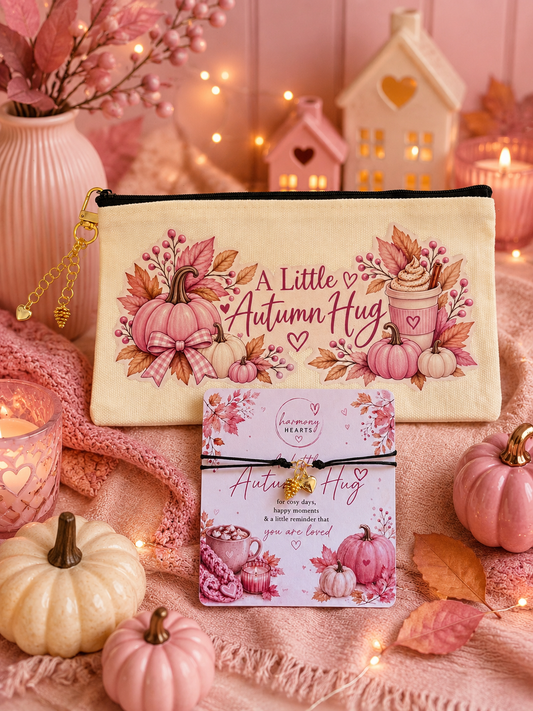 A cream fabric zip pouch printed with pink autumn pumpkins, leaves, and the phrase 'A Little Autumn Hug' is displayed with a matching greeting card with black cord bracelet attached. The scene is styled with pink knit blankets, small decorative pumpkins, string lights, and a cozy house-shaped candle holder in the background.
