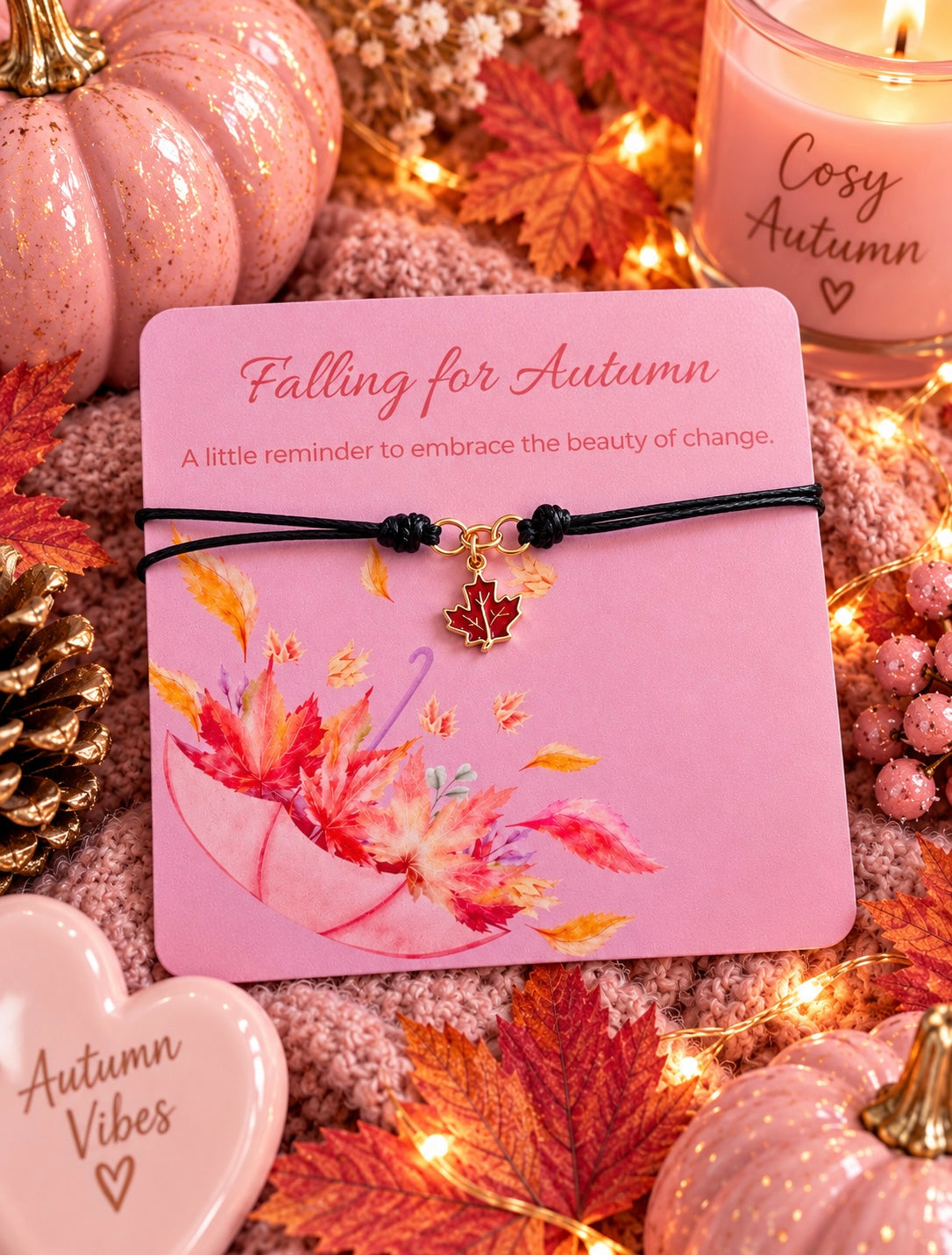 A gold maple leaf charm on a black cord bracelet, presented on a pink 'Falling for Autumn' gift card. The card is styled in an autumn-themed setting with glittery pumpkins, fall leaves, a candle, and a heart-shaped cookie.