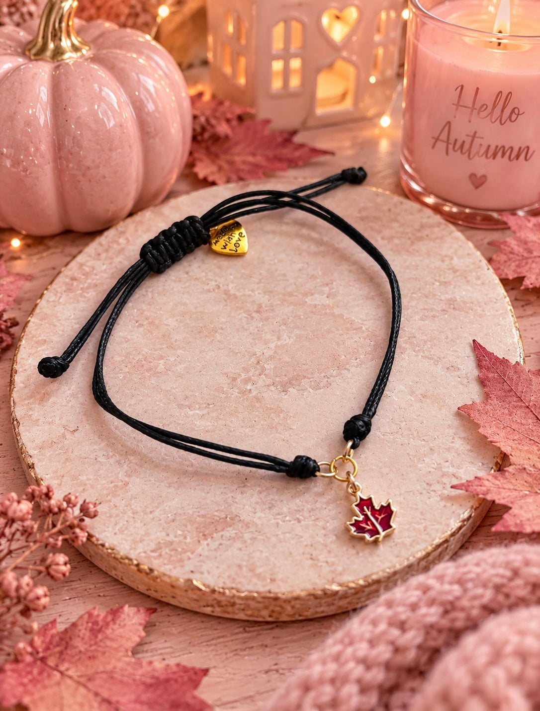 A black cord bracelet with an adjustable knot and a red maple leaf charm with gold detailing, displayed on a round stone tray with autumn-themed props including a pink pumpkin, candle, and leaves.