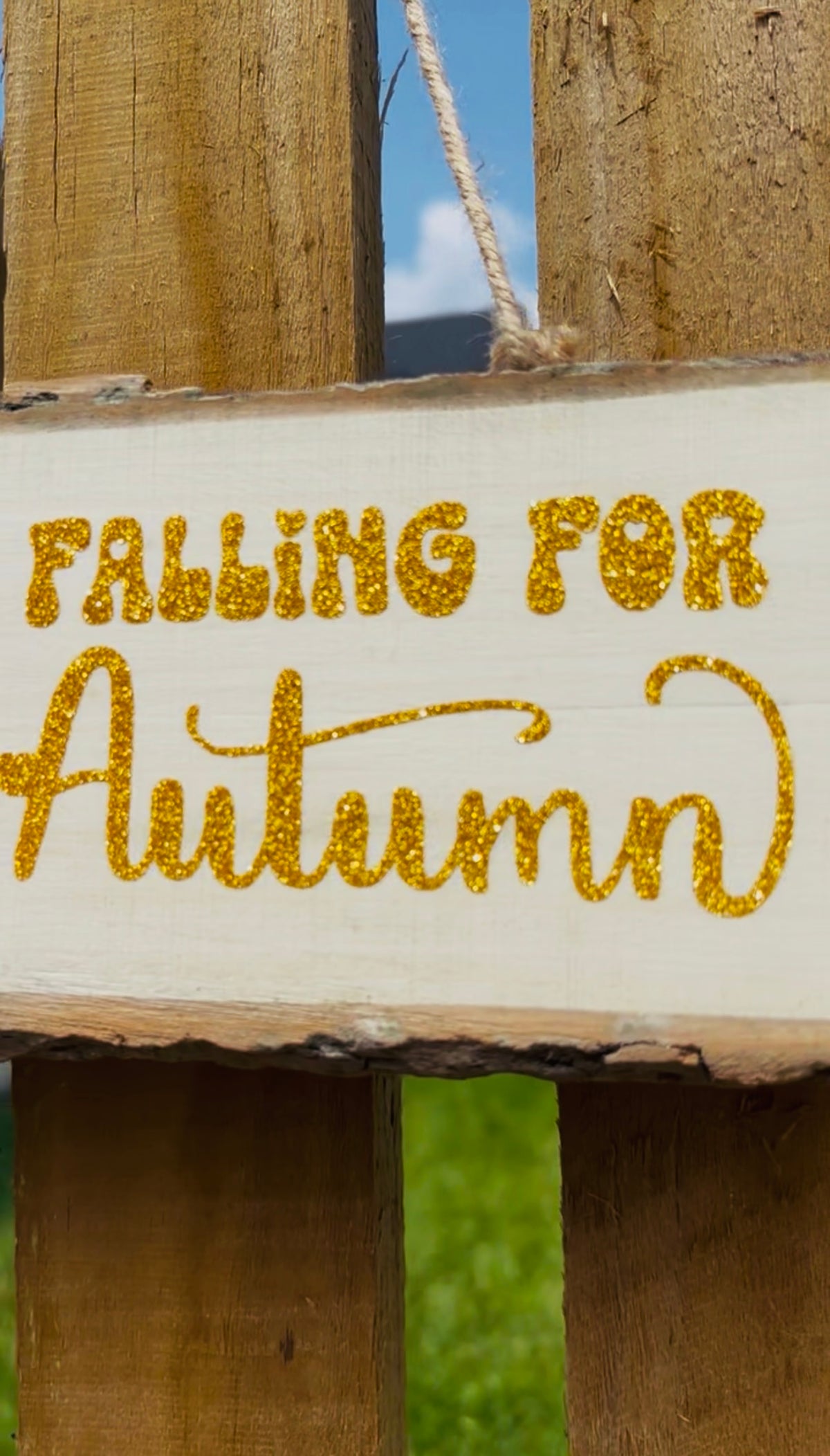 A rustic white wooden sign with the phrase 'FALLING FOR Autumn' in glittery gold lettering, hanging against a weathered wooden fence with a blurred outdoor background.