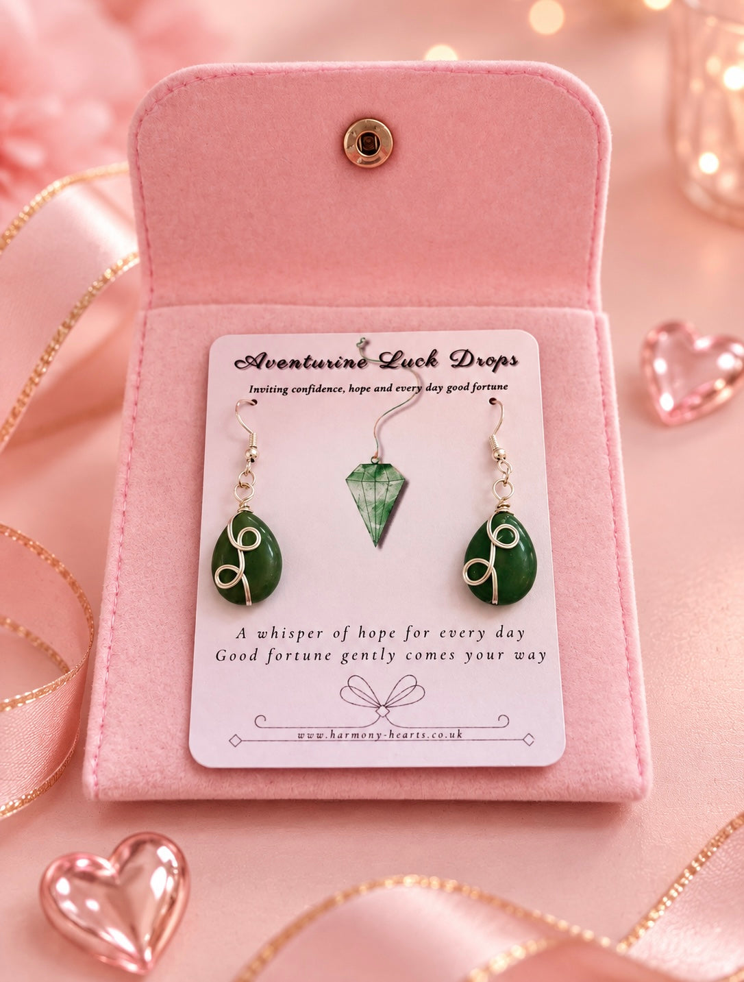 A pair of green gemstone drop earrings with silver wire wrapping, presented on a white card insert inside an open pink velvet pouch. The display is styled on a pink background with gold ribbon and heart-shaped props.