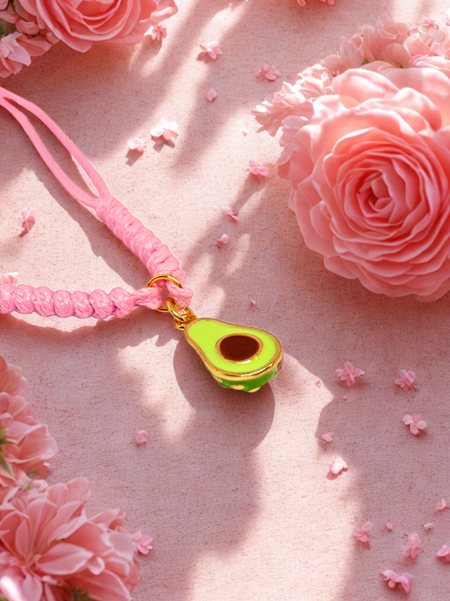 Pink braided bracelet with a green and gold avocado charm on a pink floral background