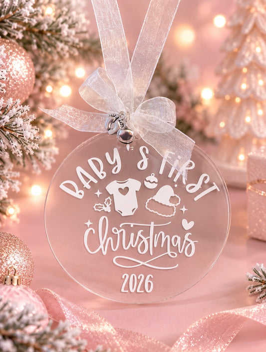 A round clear acrylic Christmas ornament with a white ribbon loop, featuring the text 'BABY'S FIRST Christmas 2026' with small baby and holiday graphics. The ornament is displayed in a festive setting with pink glitter ornaments, a flocked pine tree, and warm bokeh lights in the background.