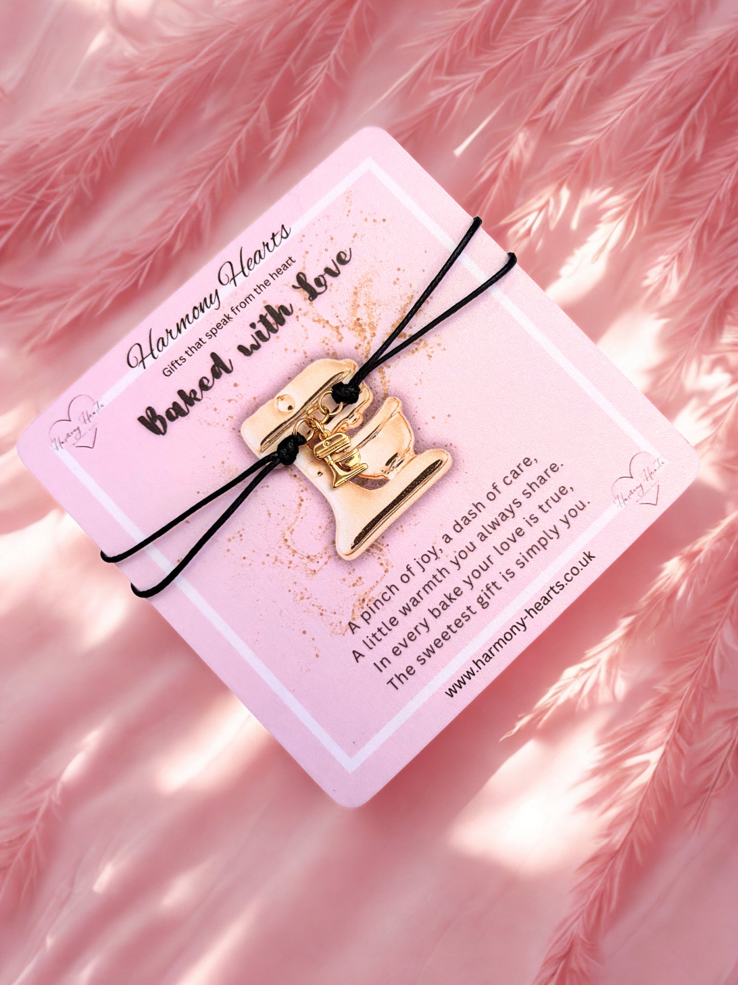 Gold food mixer charm on a pink card with text, against a pink feather background
