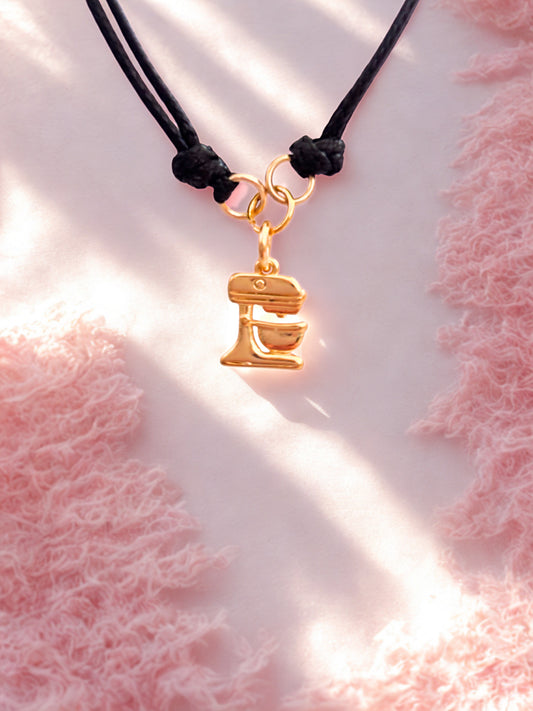 Gold food mixer charm on a black cord against a pink textured background