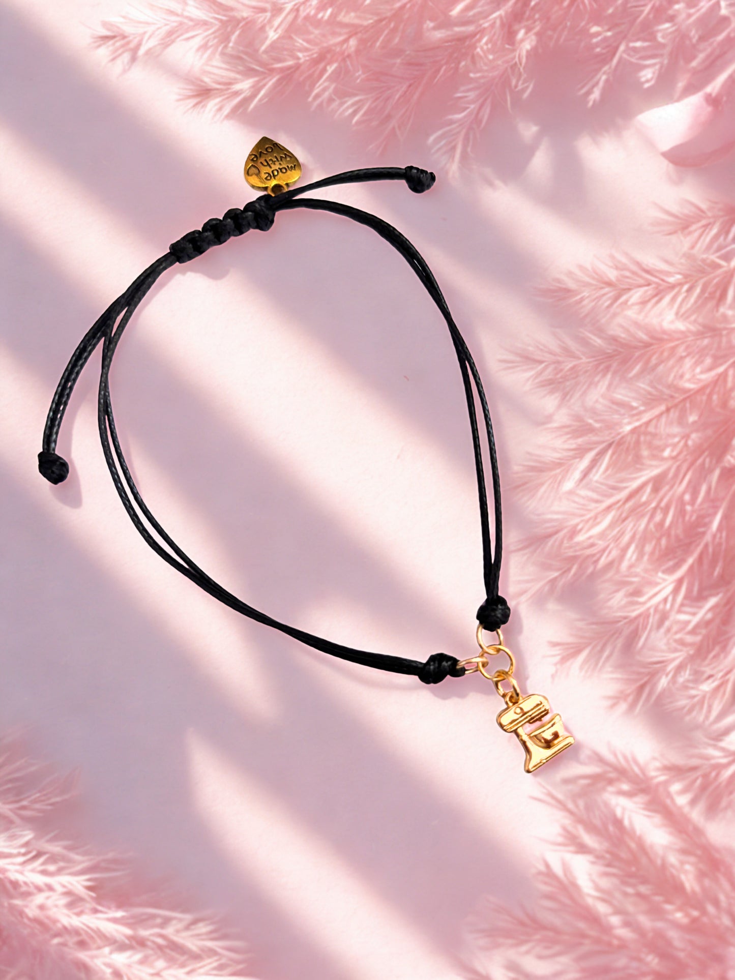Black cord bracelet with gold food mixer charm on a pink textured background