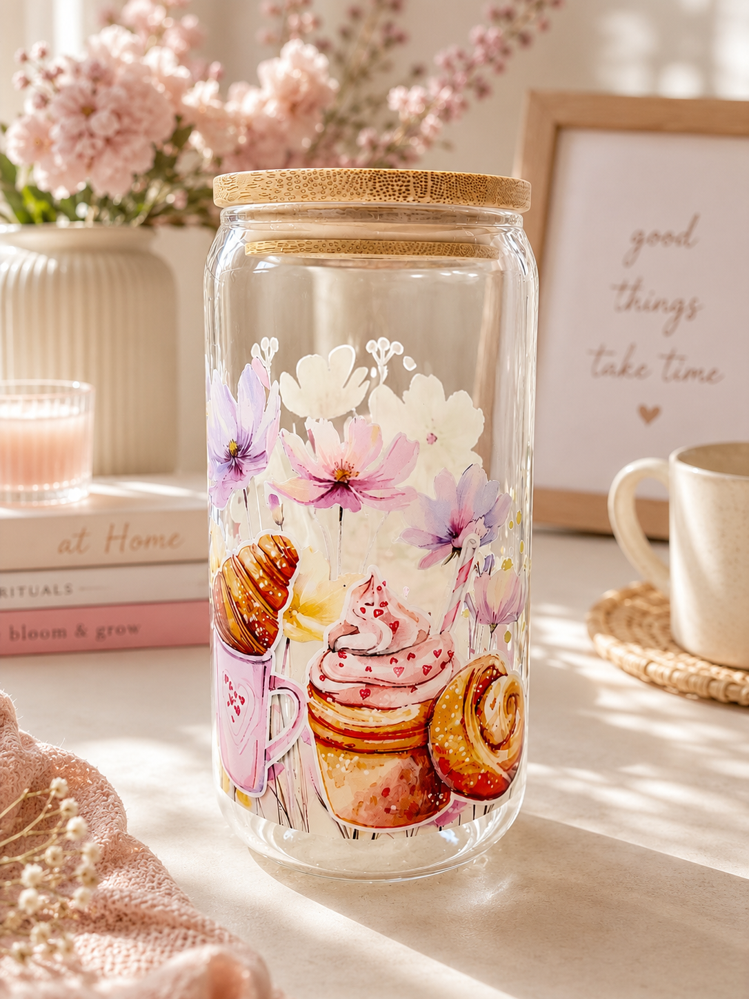 A clear glass cup with a bamboo lid, decorated with a colourful illustration of pastries, flowers, and a pink cupcake. The jar is styled on a table with a mug, a candle, and a framed print in a warm, sunlit room.