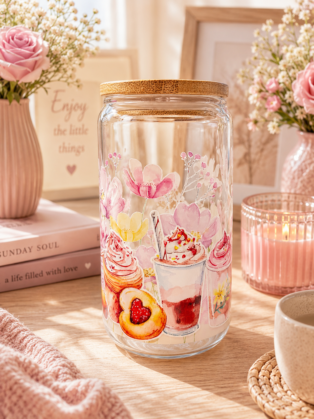 A clear glass cup with a bamboo lid, decorated with pink and yellow floral and dessert illustrations, set on a wooden table surrounded by a pink candle, flowers, and books.