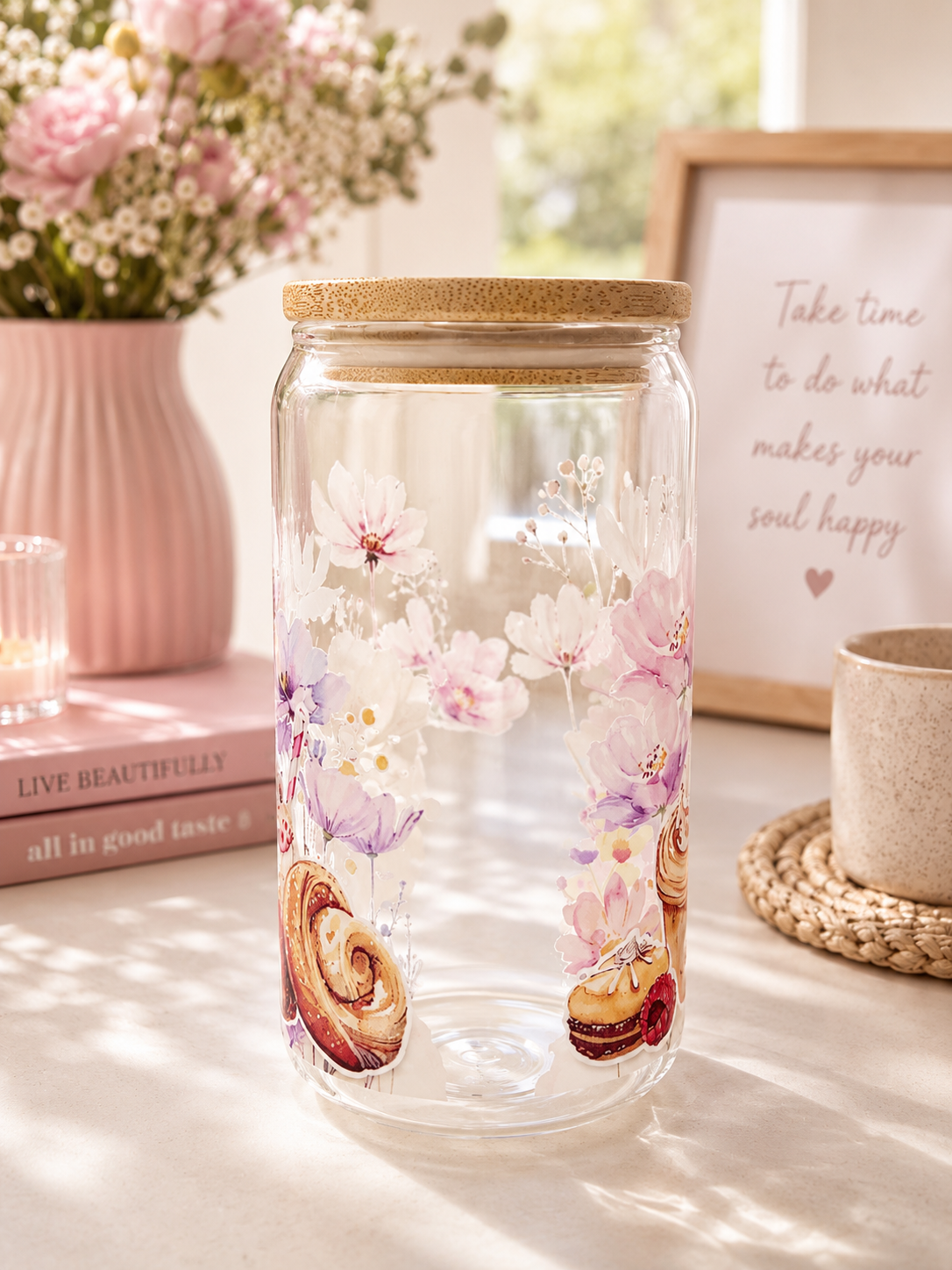 A clear glass cup with a cork lid, decorated with pink and purple cherry blossoms and pastry illustrations, set on a sunlit tabletop with books, flowers, and a framed sign in the background.