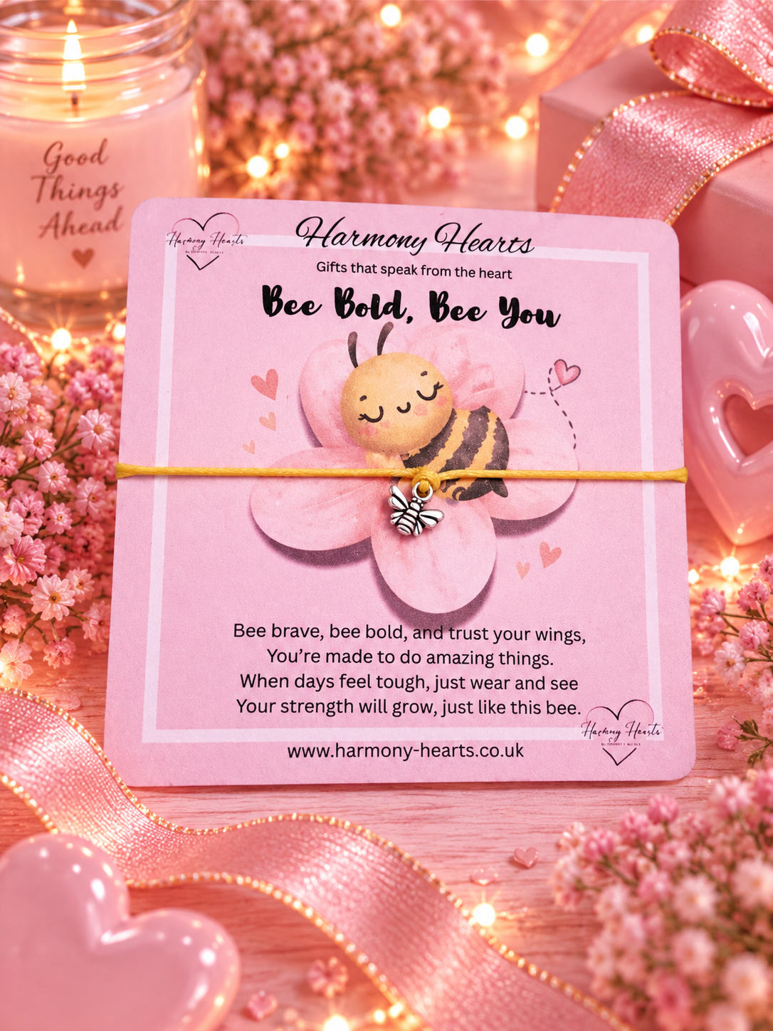 A pink Harmony Hearts card featuring a cute cartoon bee on a flower, with a bee charm on a yellow cord bracelet. The card is styled in a romantic setting with pink ribbons, heart-shaped chocolates, and a lit candle in the background.