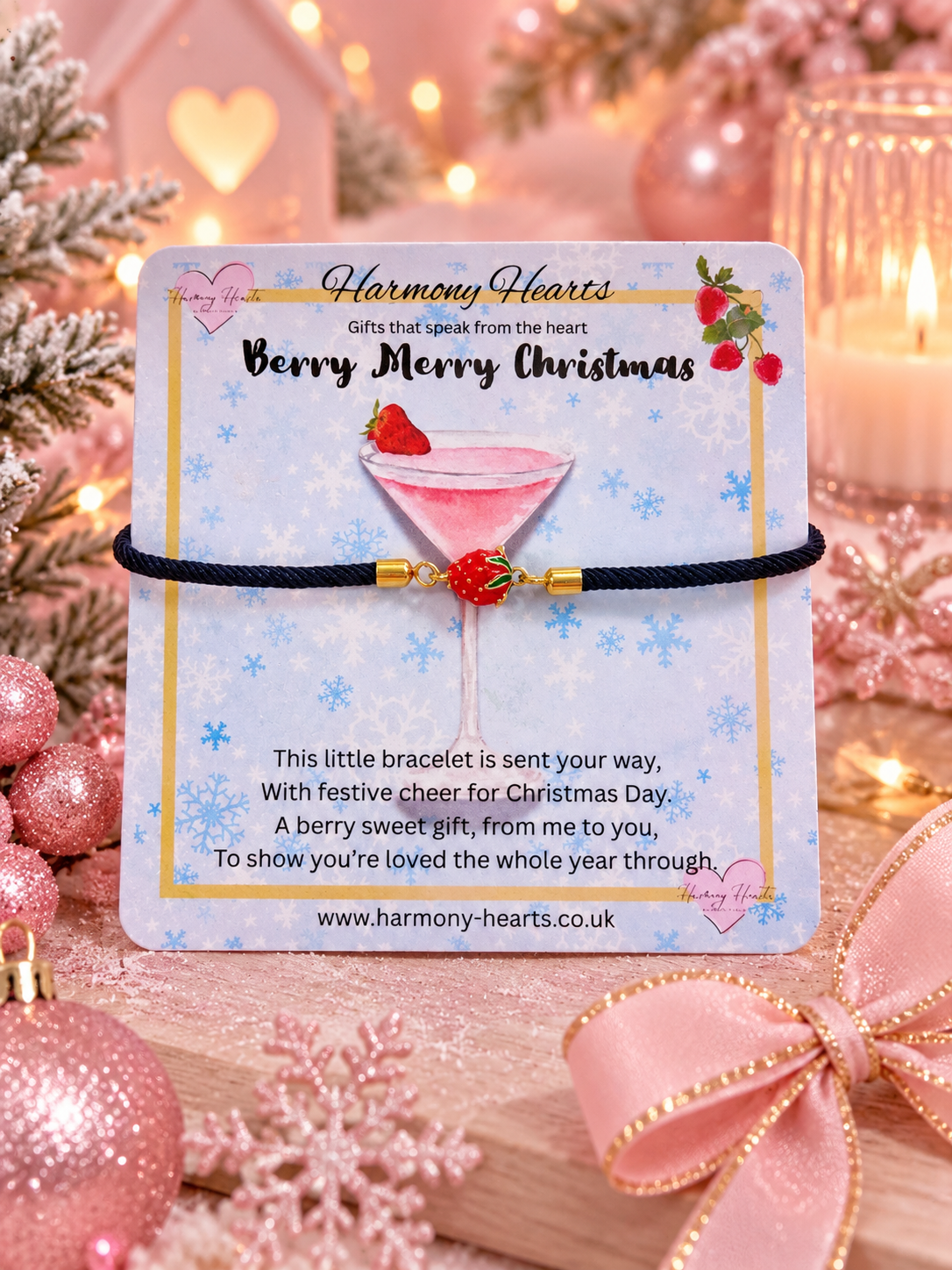 A small black cord bracelet with gold beads and a red strawberry charm displayed on a printed card reading 'Harmony Hearts Berry Merry Christmas.' The card is photographed in a festive pink holiday setting with ornaments, lights, and a ribbon.