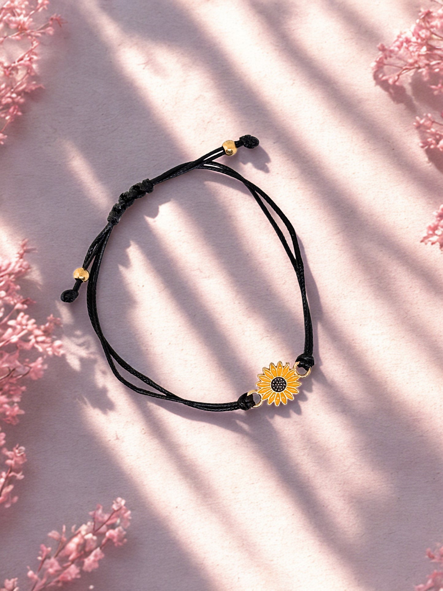Black cord bracelet with a sunflower charm on a light pink background with floral patterns