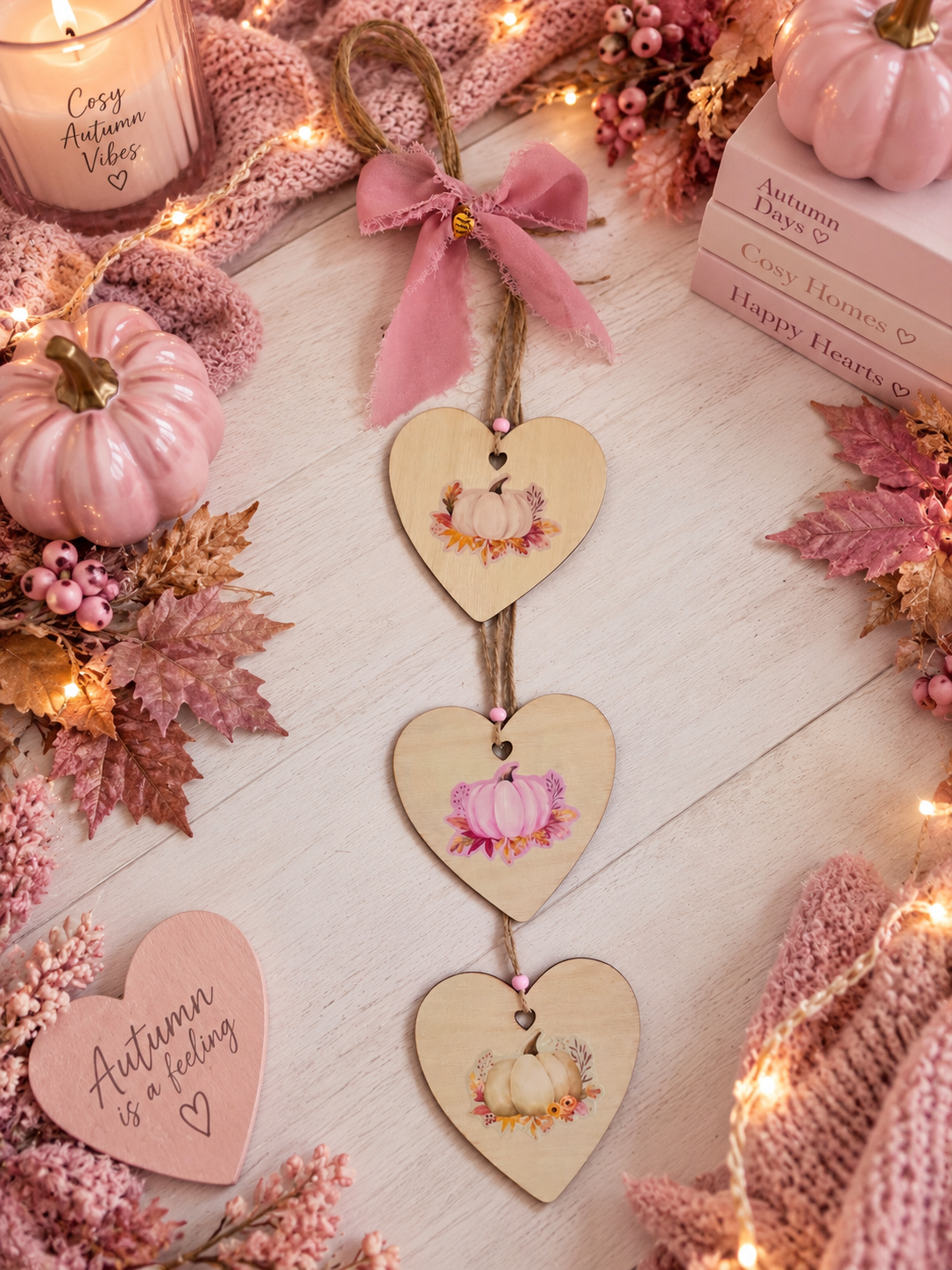 Three wooden heart-shaped hanging ornaments with pink ribbon and autumn pumpkin illustrations, styled on a light wood surface with fall decor, a candle, books, and string lights.