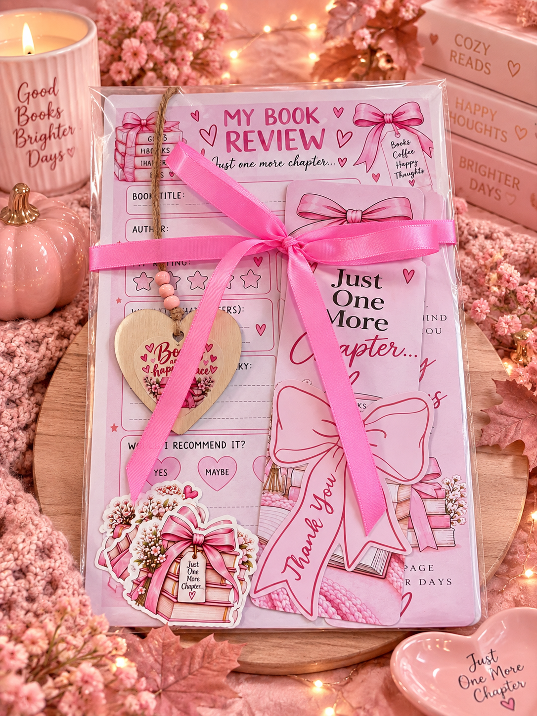 A clear plastic gift box tied with a bright pink ribbon, containing a book review stationery set with pink and white cards, stickers, and a wooden heart charm. The set is styled on a wooden board with pink books, a lit candle, and fairy lights in the background.