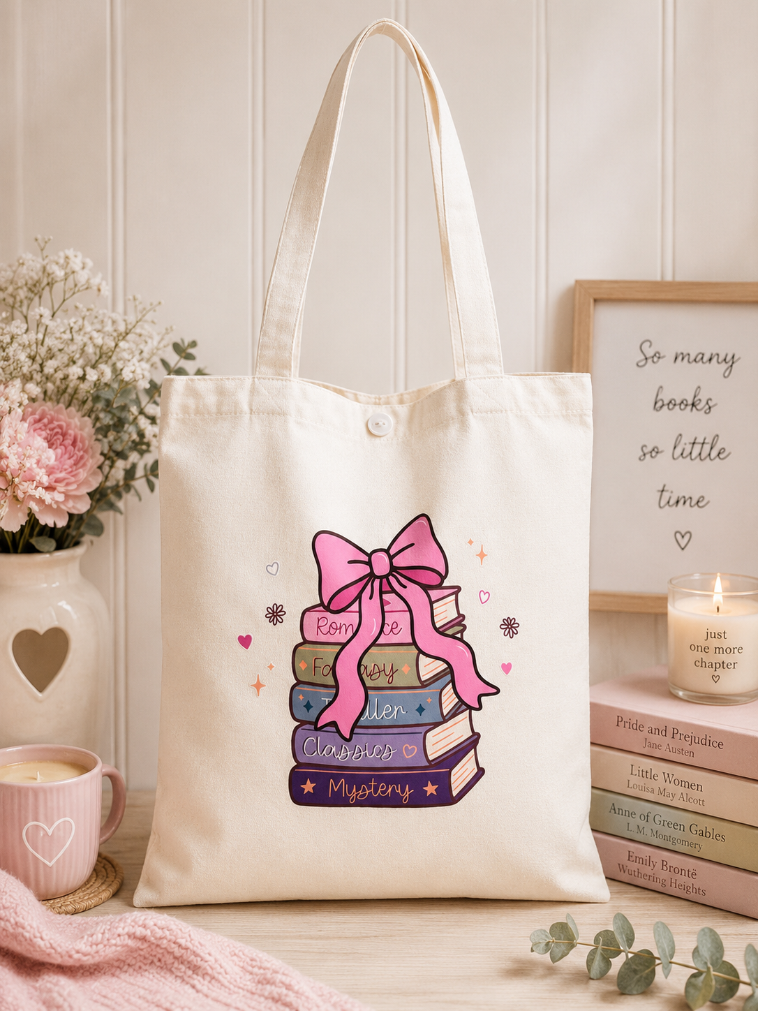 A cream canvas tote bag with a pink and purple book stack graphic, styled on a wooden surface with a pink mug, flowers, a candle, and a framed quote.