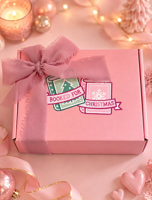 A pink gift box tied with a matching pink satin ribbon and bow, decorated with a 'Booked For Christmas' sticker. The box is set against a festive holiday background with warm lights, ornaments, and ribbons.