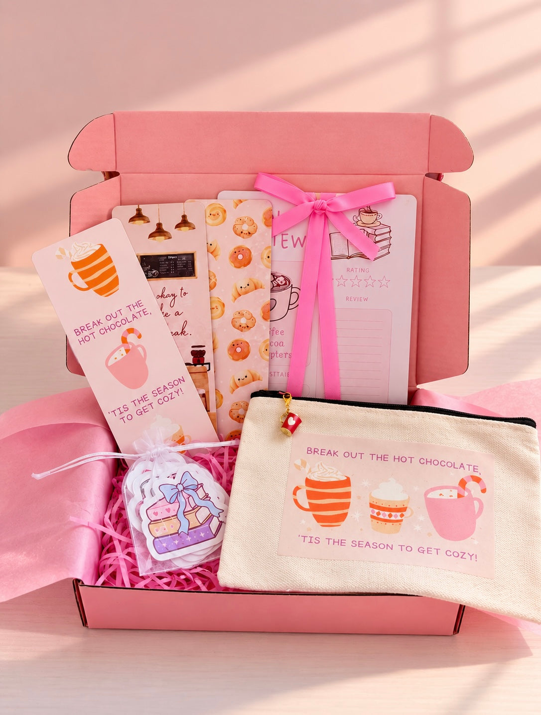 A pink gift box filled with cozy hot chocolate themed book accessories, including cards, bookmarks, a small pouch, and a decorative tag, arranged on pink paper shred.