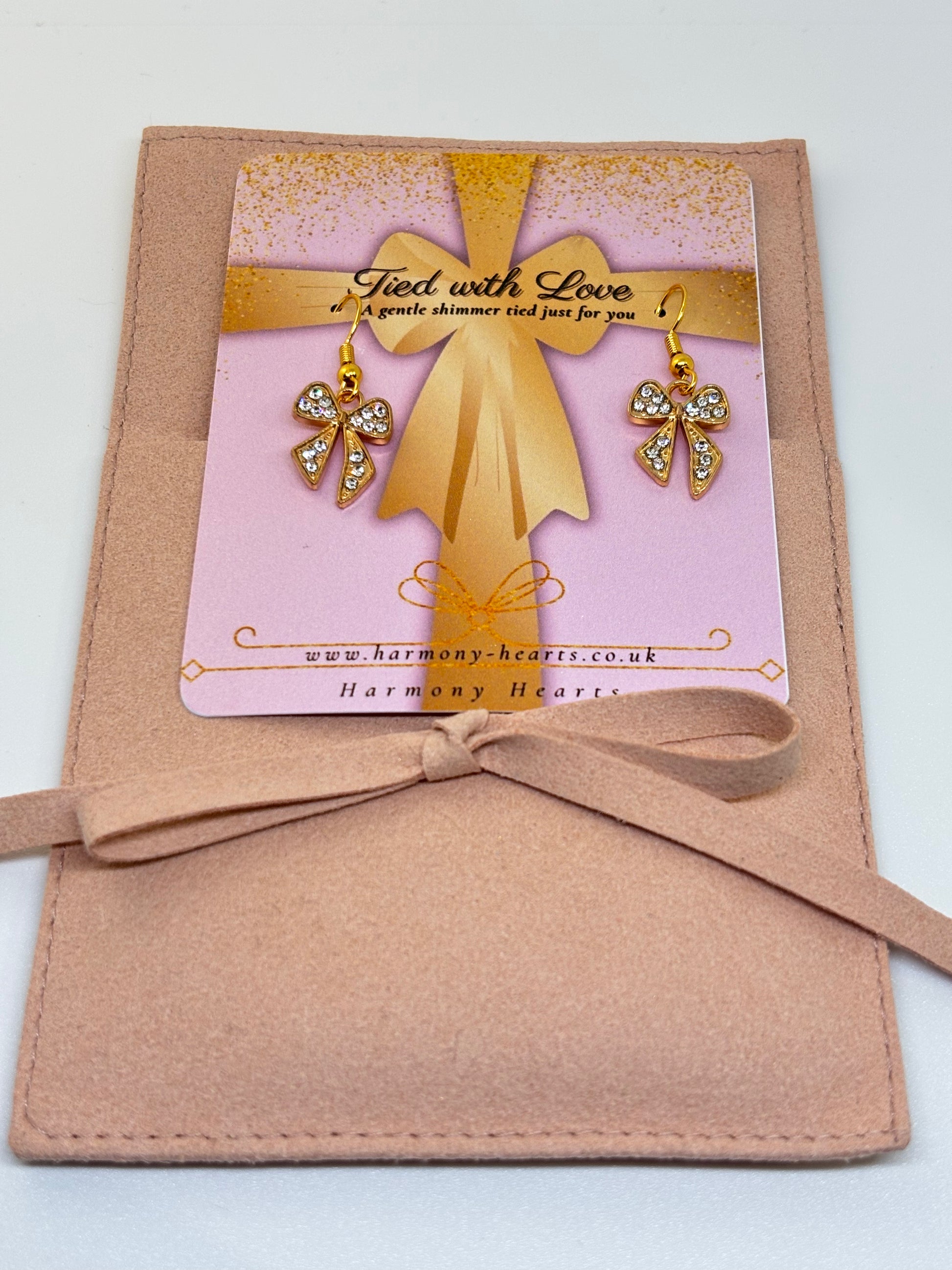 Gold bow with earrings on a pink card inside a brown envelope, labeled 'Harmony Hearts'.