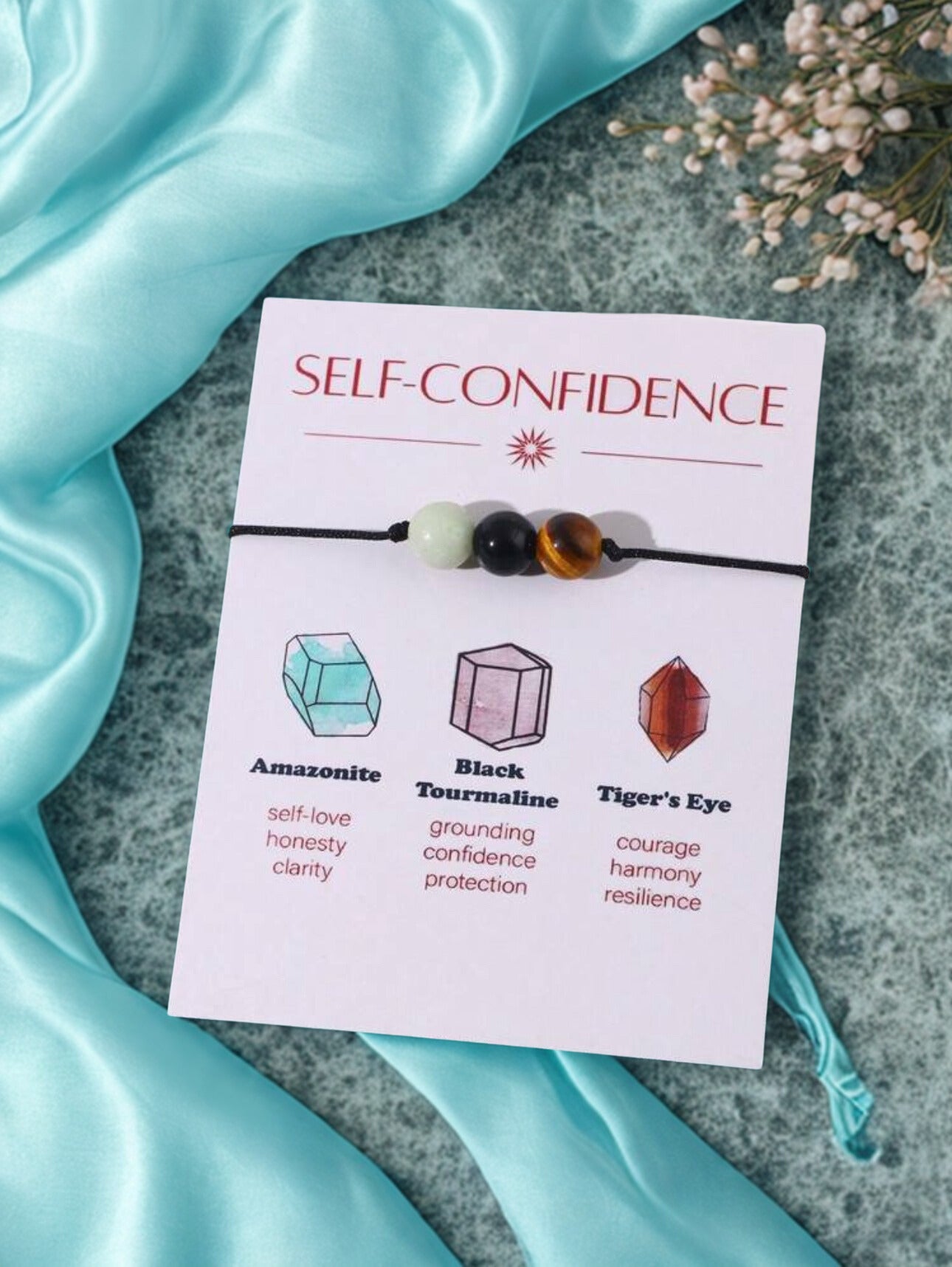 Bracelet set with 'Self-Confidence' card on a textured surface with blue fabric and flowers.