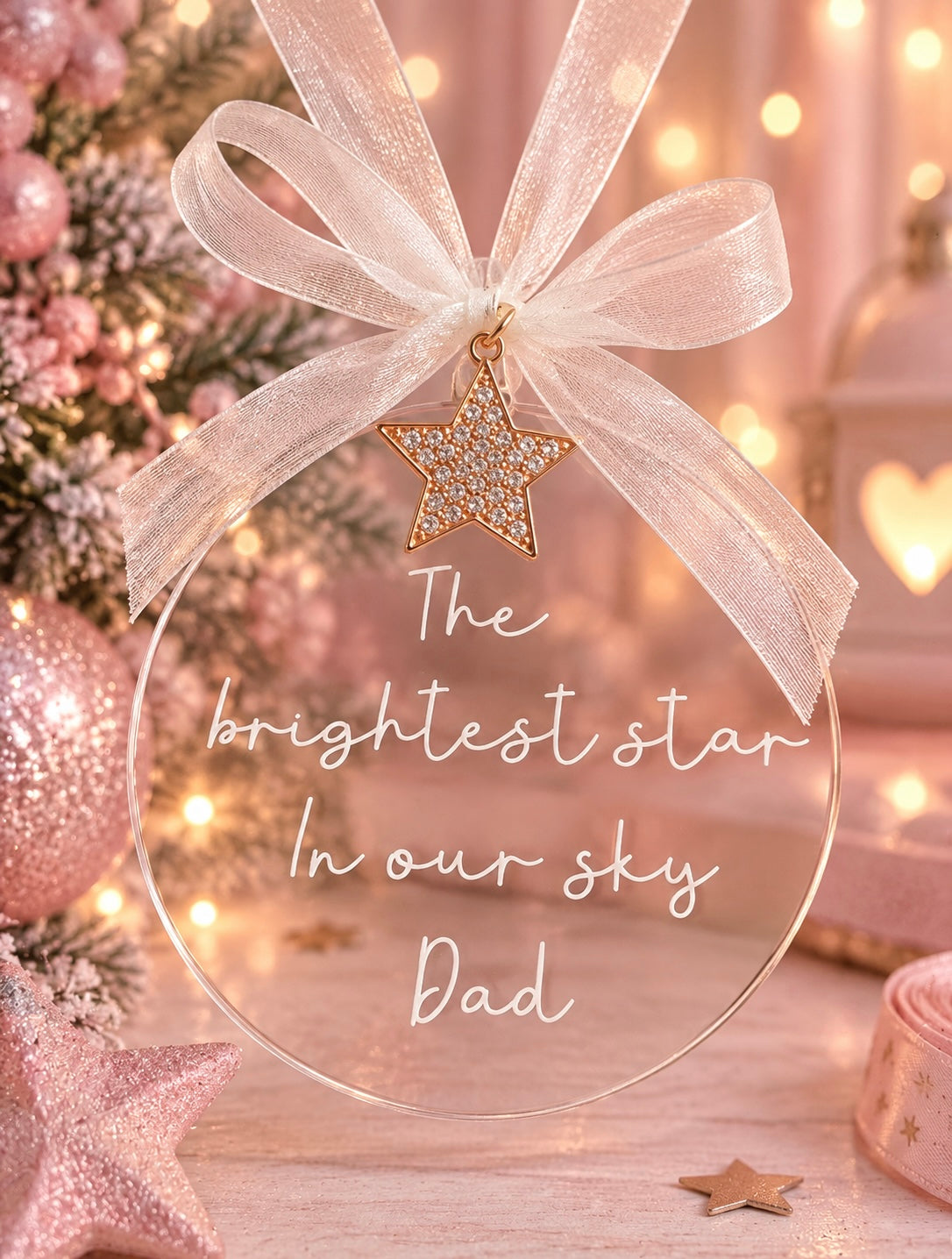A clear round hanging ornament with white script text reading 'The brightest star In our sky Dad,' tied with a sheer white ribbon and a gold star charm, shown in a softly lit holiday setting with pink ornaments and warm lights.