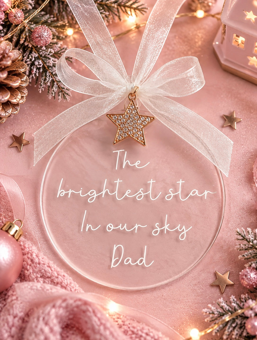 A round clear Christmas ornament with white cursive text reading 'The brightest star In our sky Dad,' tied with a sheer white ribbon and a gold star charm. The ornament is styled on a pink fabric surface with holiday decorations, stars, and warm string lights.